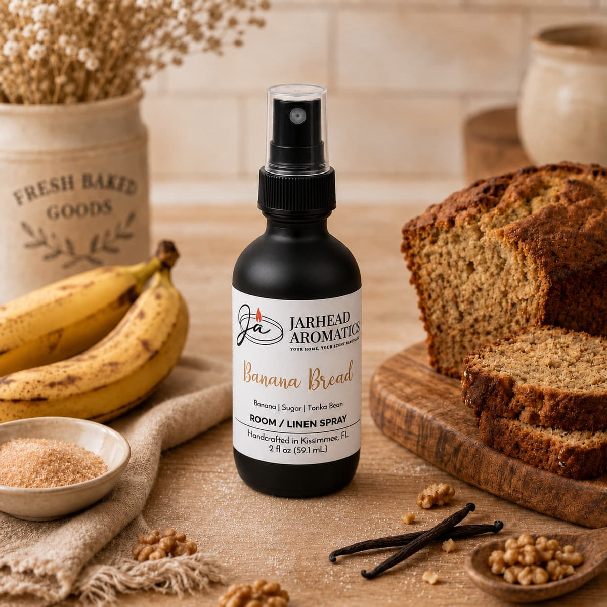 A bottle of Jarhead Aromatics Banana Bread Room & Linen Spray | Banana, Walnut & Warm Vanilla sits on a table with bananas, a sliced loaf of banana bread, a bowl of sugar, walnuts, and a basket labeled “Fresh Baked Goods” in the background, inviting you to experience the warm embrace of this irresistible home and linen fragrance with luscious notes of banana, walnut, and warm vanilla.
