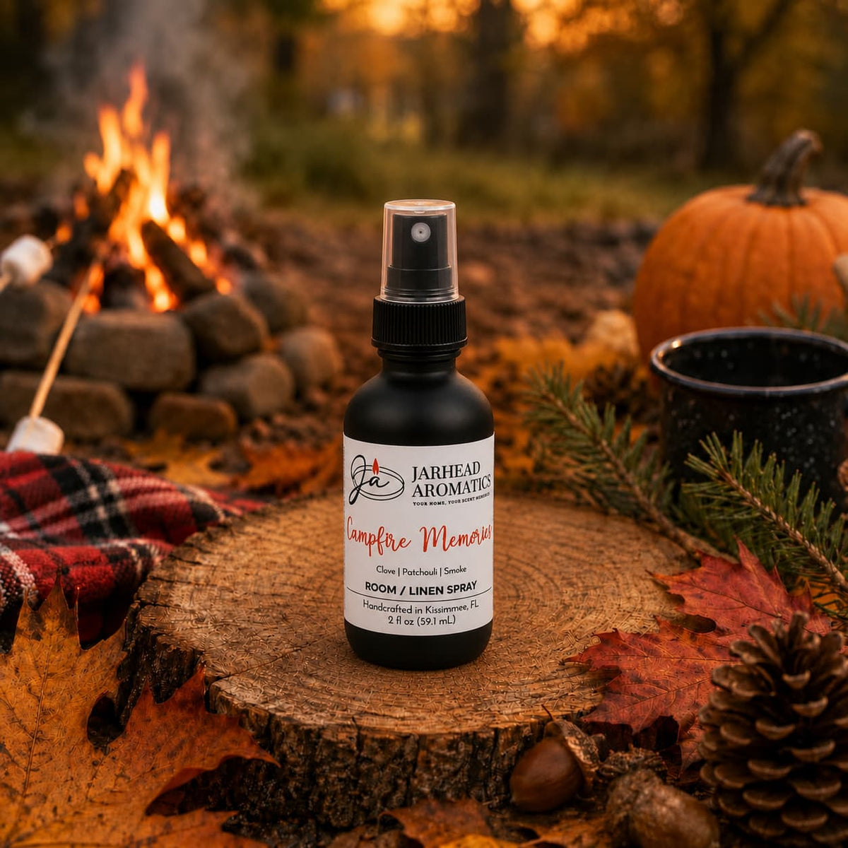 A bottle of Jarhead Aromatics Campfire Memories Room & Linen Spray (Maple, Clove & Smoky Embers) rests on a tree stump surrounded by autumn leaves, with a pumpkin, mug, plaid blanket, and crackling campfire in the background.