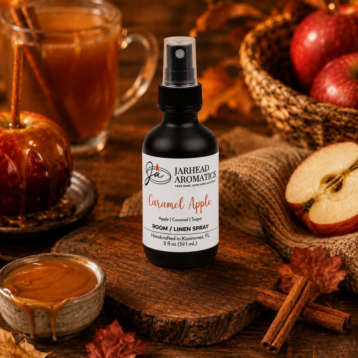 A Jarhead Aromatics Caramel Apple Room & Linen Spray | Crisp Apple, Caramel & Warm Spice bottle rests on a rustic table surrounded by caramel sauce, caramel apples, fresh apples, autumn leaves, and a mug, filling the space with its inviting fall home fragrance for a cozy seasonal atmosphere.