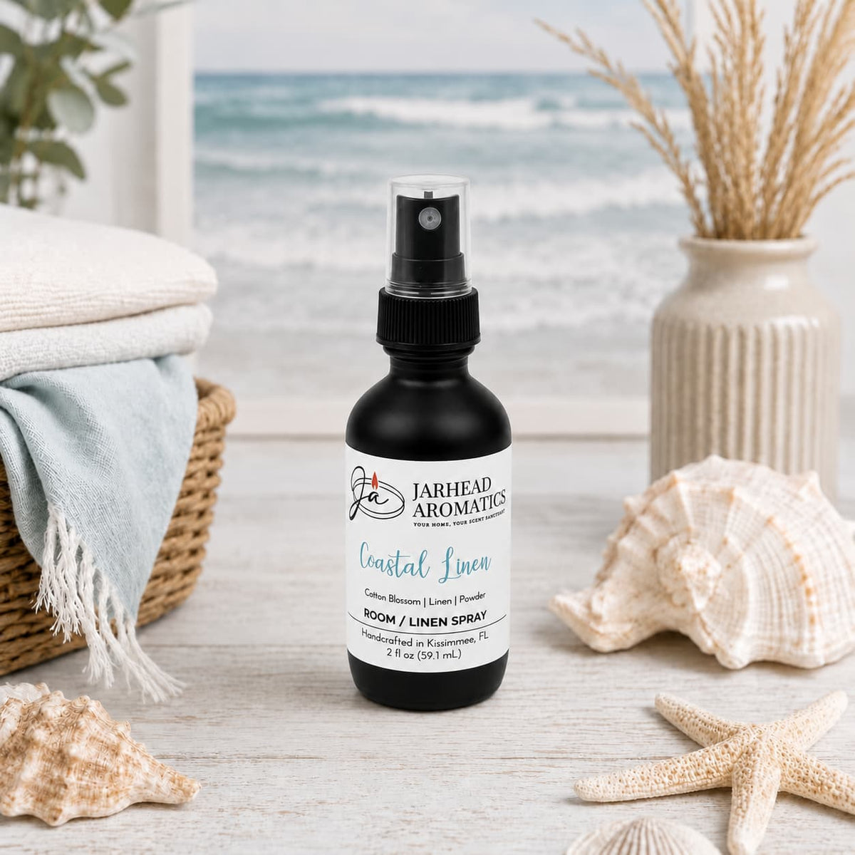 A sleek black bottle of Jarhead Aromatics’ Coastal Linen Room & Linen Spray—featuring notes of fresh linen, ozone, and sandalwood—rests atop a beach-themed table adorned with seashells, decorative vases, and neatly folded towels. The blurred ocean backdrop enhances the inviting atmosphere, evoking refreshing coastal breezes throughout your home.