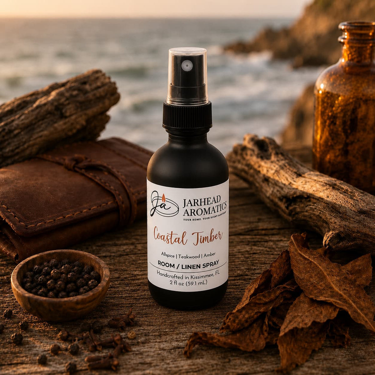 A bottle of Jarhead Aromatics Coastal Timber Room & Linen Spray | Teakwood, Leather & Warm Amber rests gracefully on driftwood, encircled by spices and dried leaves, with an ocean and rocky coastline bathed in sunset light—evoking the essence of a luxury room spray that infuses any space with a woodsy home fragrance.