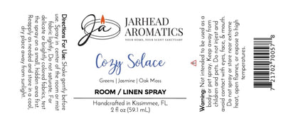 White label for Jarhead Aromatics Cozy Solace Room & Linen Spray | Fresh Greens, Jasmine & Oak Moss, 2 fl oz. This home fragrance includes directions, warnings, and scent notes of fresh greens, jasmine, and oak moss. Logo and text are black/blue with a small orange candle graphic.