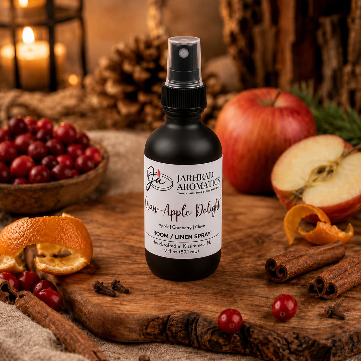 A bottle of Jarhead Aromatics Cran-Apple Delight Room & Linen Spray rests on a wooden surface, surrounded by fresh apples, cranberries, orange peels, and autumn decorations, with candles and pinecones in the background—a charming display that perfectly embodies the cozy scent of cranberry, apple, and warm cinnamon in this inviting home fragrance.
