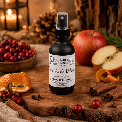 A bottle of Jarhead Aromatics Cran-Apple Delight Room & Linen Spray rests on a wooden surface, surrounded by fresh apples, cranberries, orange peels, and autumn decorations, with candles and pinecones in the background—a charming display that perfectly embodies the cozy scent of cranberry, apple, and warm cinnamon in this inviting home fragrance.