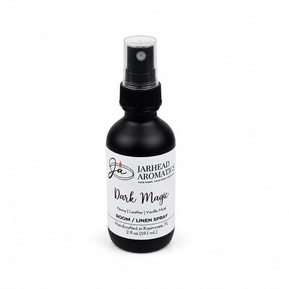 A black spray bottle labeled Jarhead Aromatics Dark Magic Room & Linen Spray is displayed on a white background, featuring an enchanting home fragrance with notes of peach, plum, dark florals, and vanilla musk; 2 oz (59 ml), handcrafted in Kissimmee, FL.