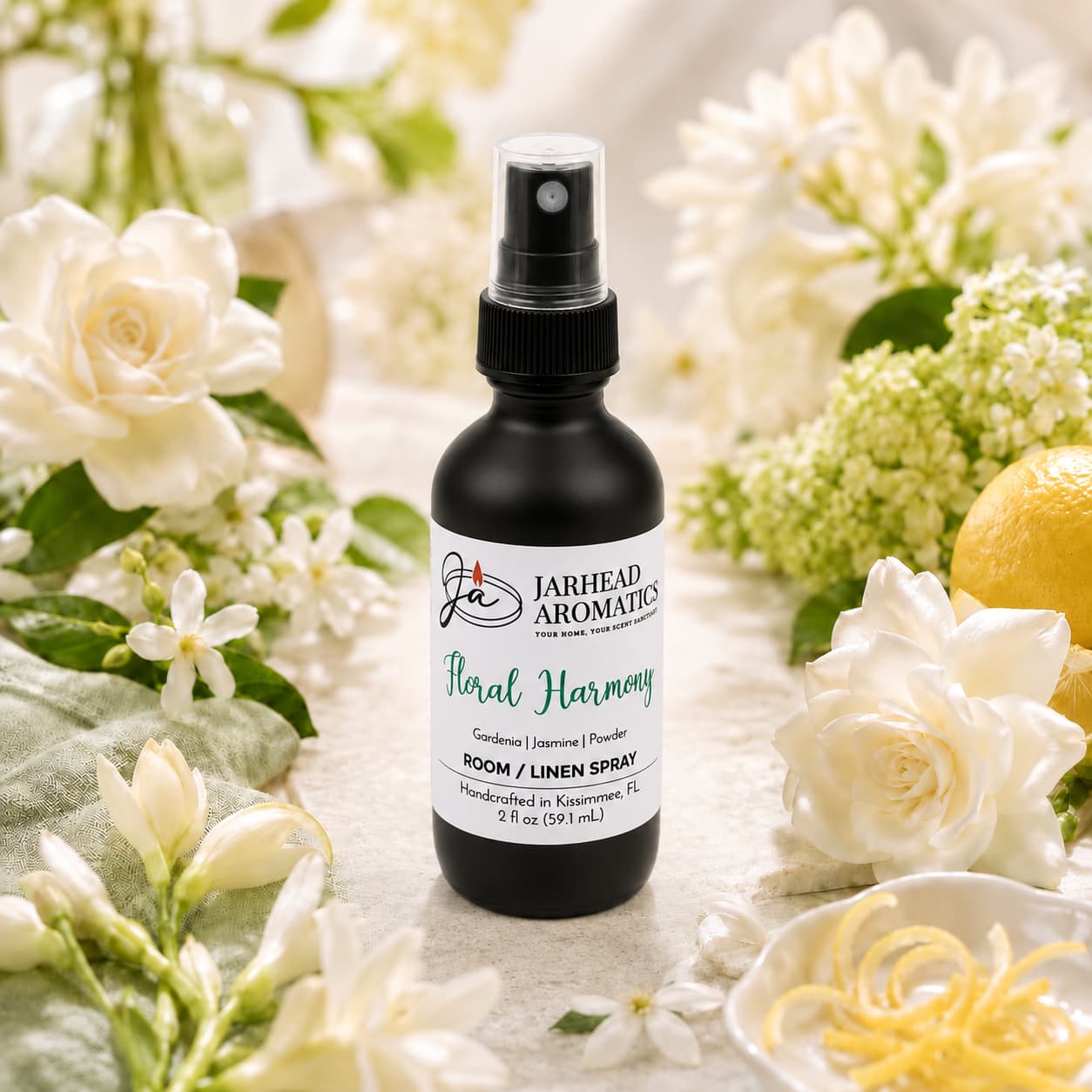 A bottle of Jarhead Aromatics Floral Harmony Room & Linen Spray | Gardenia, Jasmine & Tuberose rests among white flowers, green leaves, and a lemon on a light-colored surface, creating a fresh and elegant scene.