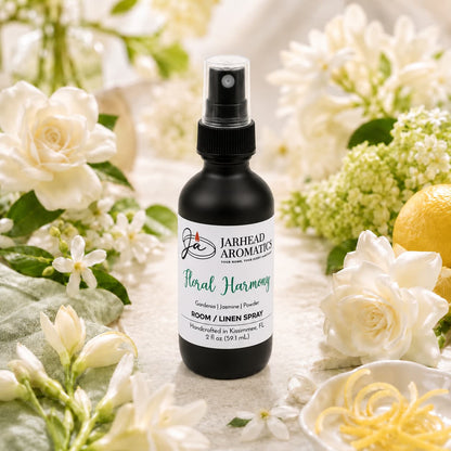 A bottle of Jarhead Aromatics Floral Harmony Room & Linen Spray | Gardenia, Jasmine & Tuberose rests among white flowers, green leaves, and a lemon on a light-colored surface, creating a fresh and elegant scene.
