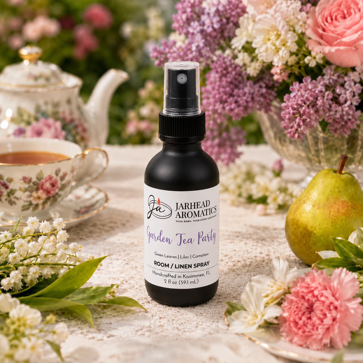 A bottle of Jarhead Aromatics Garden Tea Party Room & Linen Spray in Rose, Lilac & Jasmine rests on a table surrounded by flowers, a teapot, a teacup, and a pear, creating an elegant and inviting tea party scene that beautifully captures the essence of this garden-inspired fragrance.