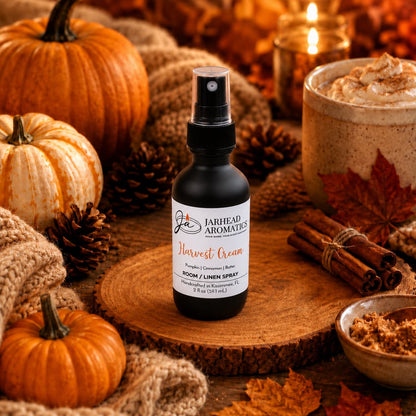 A bottle of Jarhead Aromatics Harvest Cream Room & Linen Spray | Pumpkin Spice, Brown Sugar & Vanilla Cream rests on a wooden surface amidst mini pumpkins, pinecones, cinnamon sticks, and a creamy beverage. Warm lighting and scattered fall leaves complete the scene, evoking a cozy autumn ambiance with this true gourmand home fragrance that captures the seasonal essence.