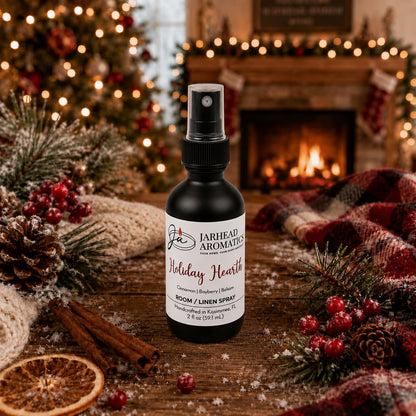 A Jarhead Aromatics Holiday Hearth Room & Linen Spray | Cinnamon Clove, Bayberry & Balsam bottle sits on a wooden table surrounded by cinnamon sticks, pine branches, and dried orange slices, with a Christmas tree and fireplace in the background.