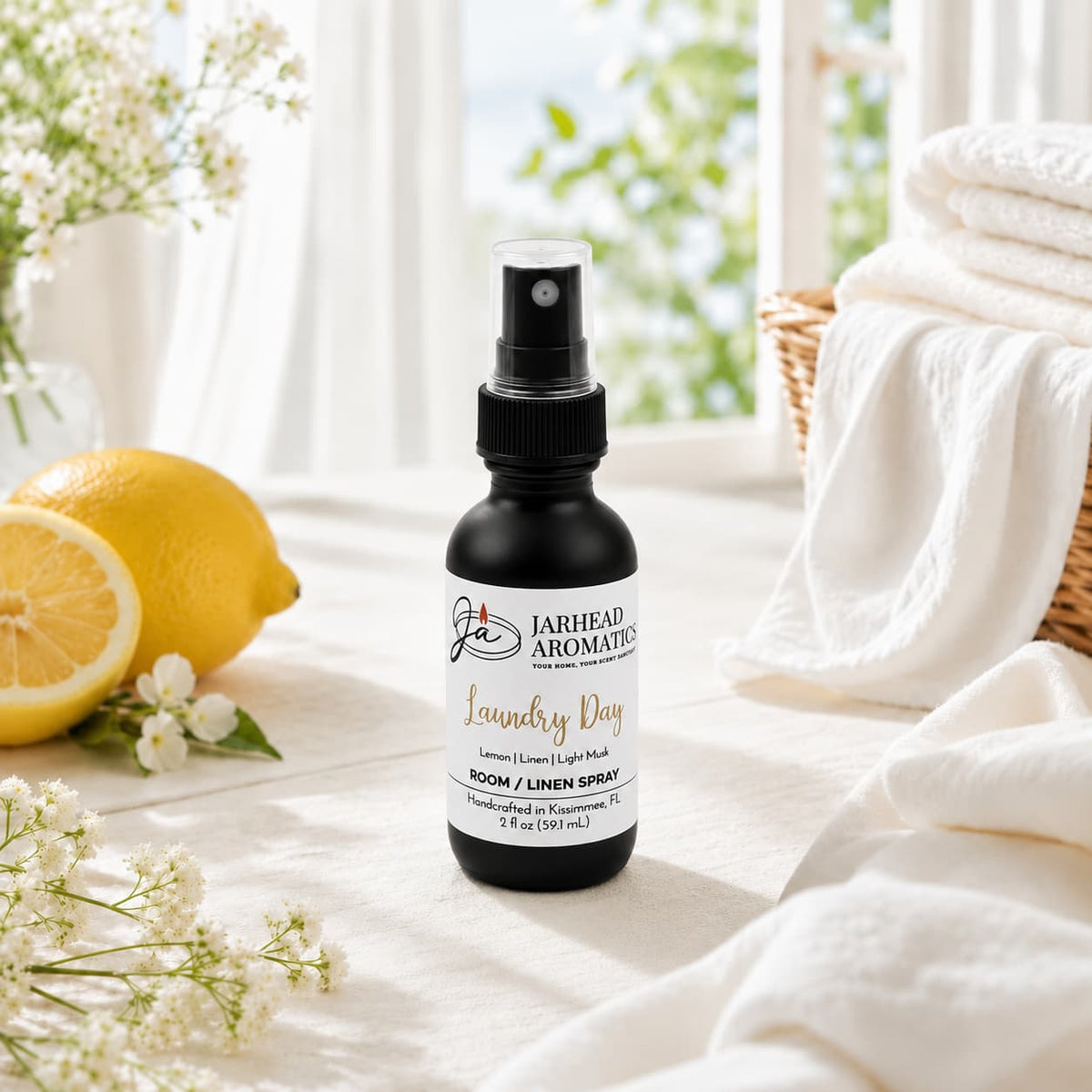 A black spray bottle of Jarhead Aromatics’ Laundry Day Room & Linen Spray | Fresh Linen, Lemon & Clean Musk rests on a table with fresh towels, a sliced lemon, flowers, and green foliage in the background, evoking the scent of a clean home with fresh linens.