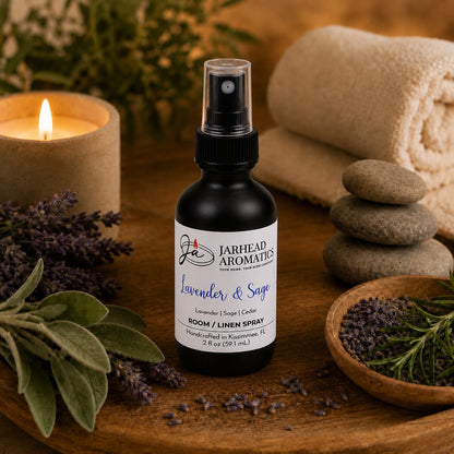 A bottle of Jarhead Aromatics Lavender & Sage Room & Linen Spray, featuring lavender, sage, and sandalwood, is displayed amidst fresh lavender, a lit candle, smooth stones, a towel, and a wooden bowl of herbs—creating a relaxing spa-inspired scene that brings calming fragrance to any space.