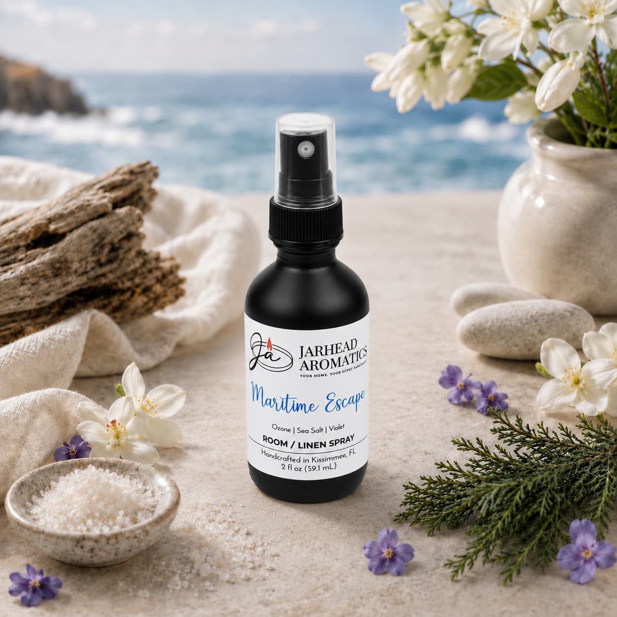 A bottle of Jarhead Aromatics Maritime Escape Room & Linen Spray | Sea Salt, Ozone & Jasmine rests on a table adorned with flowers, stones, and a bowl of salt, infusing your home fragrance collection with a refreshing coastal scent. In the softly blurred background, the ocean and cliffs evoke the tranquil ambiance of the shore.