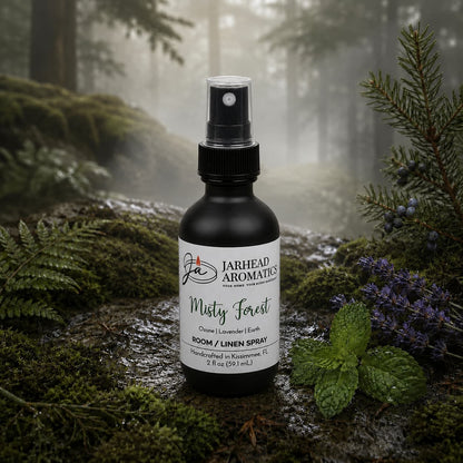 A bottle of Misty Forest Room & Linen Spray | Ozone, Moss & Juniper by Jarhead Aromatics rests on a moss-draped log in a mist-shrouded forest, encircled by lush ferns, vibrant purple blooms, and fresh greenery—a fitting scene for this nature-inspired home fragrance that captures the enchanting aroma of ozone, moss, and juniper.