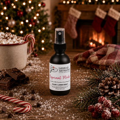 A bottle of Jarhead Aromatics Peppermint Mocha Room & Linen Spray sits on a wooden table surrounded by a mug of hot cocoa, chocolate pieces, a candy cane, and a pine branch.
