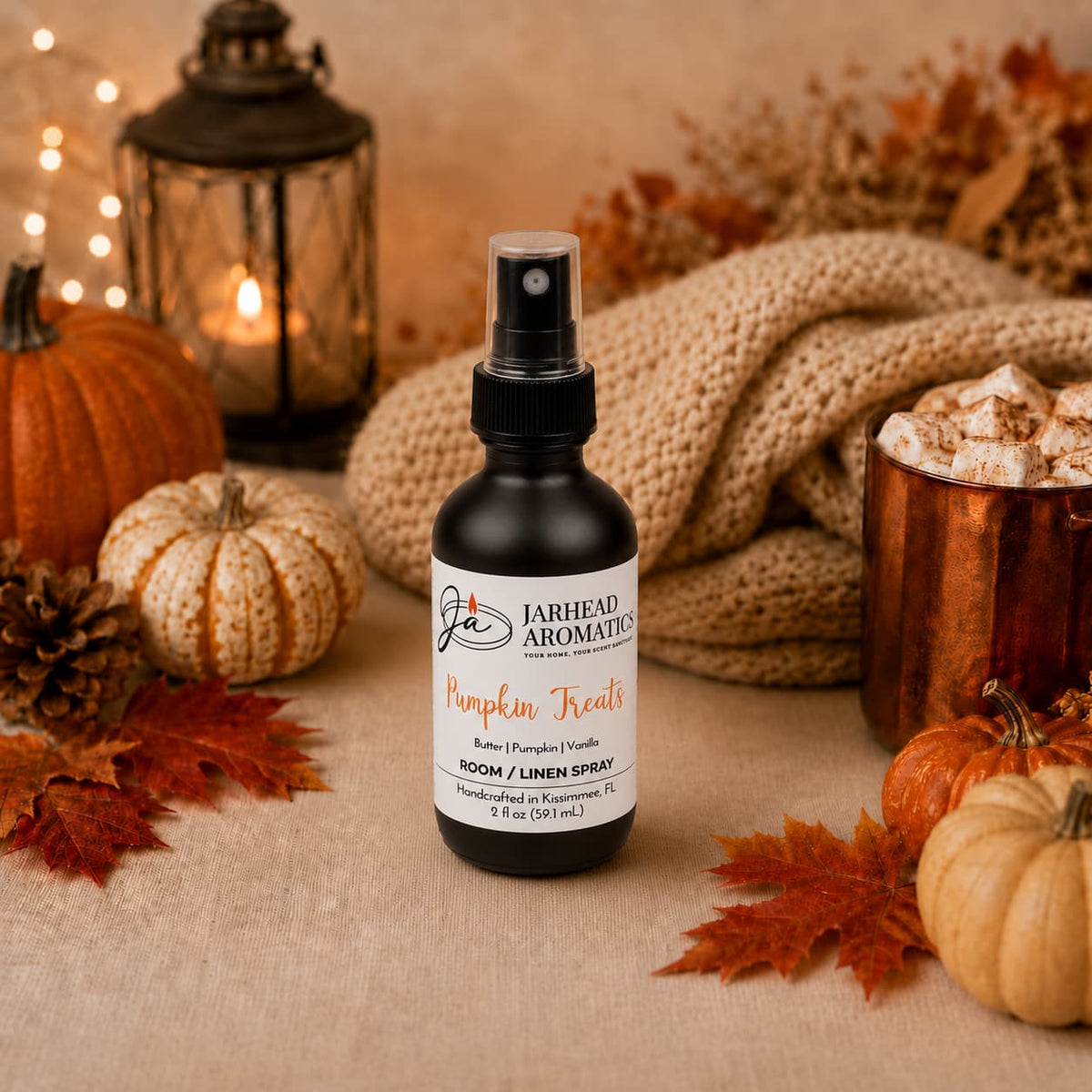 A bottle of Jarhead Aromatics’ Pumpkin Treats Room & Linen Spray—featuring notes of pumpkin, caramel, and maple pecan—rests on a table surrounded by small pumpkins, autumn leaves, a lantern, a beige knit blanket, and a mug of marshmallows. The inviting fall fragrance fills the air and transforms the space into the perfect cozy autumn scene.