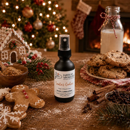 A bottle of Santa’s Cookies Room & Linen Spray | Ginger, Brown Sugar, Molasses & Vanilla sits on a wooden table surrounded by gingerbread men, chocolate chip cookies, a glass of milk, and Christmas decorations.