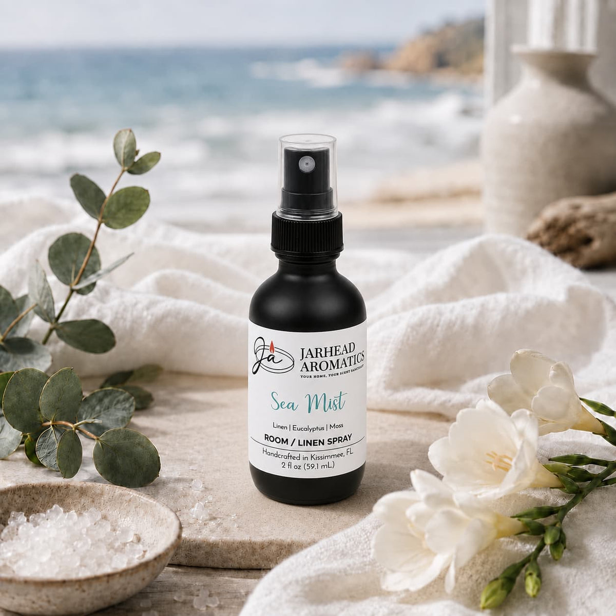 A bottle of Jarhead Aromatics Sea Mist Room & Linen Spray | Fresh Linen, Sea Salt & Eucalyptus rests on a towel alongside eucalyptus, white flowers, sea salt, and vases, with a softly blurred ocean and beach in the background—evoking the fresh coastal essence of this home fragrance for your spa-inspired room spray experience.