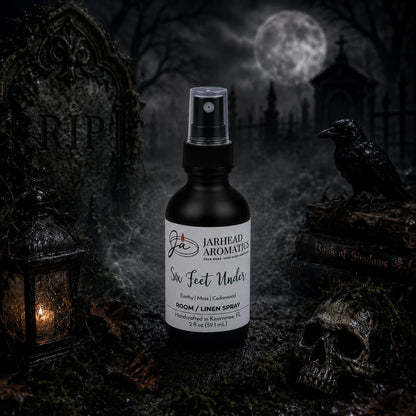 A bottle of Jarhead Aromatics’ Six Feet Under Room & Linen Spray | Damp Earth, Moss & Ancient Oak stands in a foggy graveyard surrounded by tombstones, a raven, a skull, and the Book of Shadows beneath a dark, eerie sky. The lingering aroma of damp earth, moss, and ancient oak drifts through the air—an enchanting home fragrance designed for those who relish the mysterious and macabre.