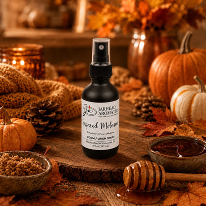 A bottle of Jarhead Aromatics Sugared Molasses Room & Linen Spray—featuring notes of buttercream, honey, and dark molasses—rests atop a rustic table surrounded by honey, brown sugar, pumpkins, pinecones, and autumn leaves in a warmly lit fall setting, perfect for filling your home with the comforting aroma of this inviting gourmand fragrance.