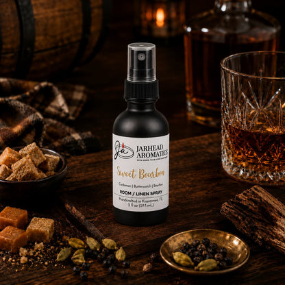 A bottle of Jarhead Aromatics Sweet Bourbon Room & Linen Spray | Bourbon, Butterscotch & Brown Sugar rests on a wooden table surrounded by sugar cubes, spices, a glass of whiskey, and a whiskey barrel in the background, embodying an inviting gourmand fragrance that elevates your home scent experience.