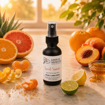 A bottle of Jarhead Aromatics Sweet Sunrise Room & Linen Spray | Lemon, Orange, Peach & Citrus rests on a marble surface surrounded by vibrant citrus fruits and hints of honey and sugar, as sunlight pours through a nearby window—perfectly capturing the uplifting essence of this citrus home fragrance to enliven any room.