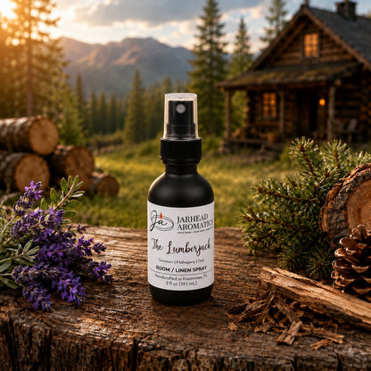 A bottle of The Lumberjack Room & Linen Spray by Jarhead Aromatics—featuring notes of mahogany, cedarwood, and oak—rests on a wooden surface outdoors, nestled among pinecones and lavender, with a log cabin and trees bathed in sunset light in the background, perfectly capturing the essence of a woodsy home fragrance in every spritz.