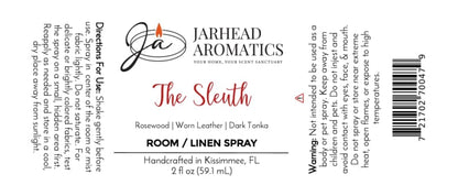 A Jarhead Aromatics The Sleuth Room & Linen Spray bottle, featuring an elegant white label with directions, warnings, and company details, showcases enticing notes of leather, rosewood, and sandalwood—ideal for enhancing your home fragrance experience.