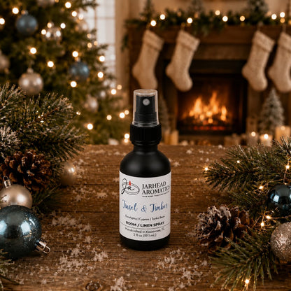 A bottle of Jarhead Aromatics Tinsel & Timber Room & Linen Spray sits on a wooden surface with pine branches, ornaments, and artificial snow, set against a Christmas tree and fireplace.