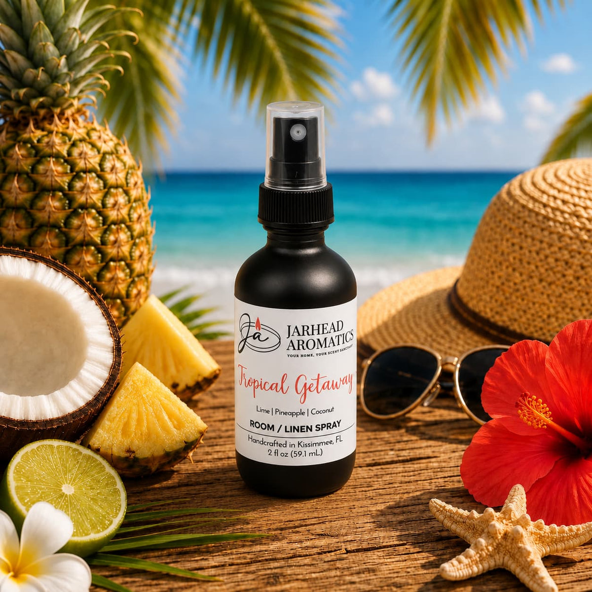 A bottle of Jarhead Aromatics Tropical Getaway Room & Linen Spray | Coconut, Mango & Pineapple rests on the beach among pineapples, sunglasses, a straw hat, and a red hibiscus flower, infusing the air with an inviting home fragrance while the ocean and blue sky create a stunning backdrop.