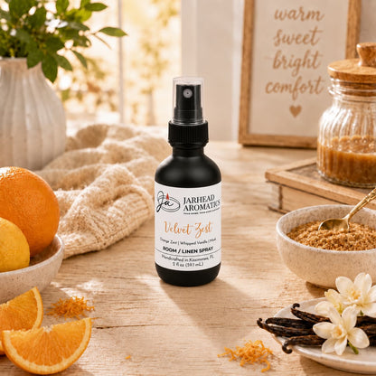 A spray bottle of Jarhead Aromatics Velvet Zest Room & Linen Spray | Citrus, Vanilla & Brown Sugar rests on a wooden table surrounded by oranges, vanilla beans, brown sugar, and white flowers. In the background, a sign reading “warm sweet bright comfort” highlights the vibrant display—perfectly reflecting this citrus-vanilla fragrance’s inviting charm for any home.