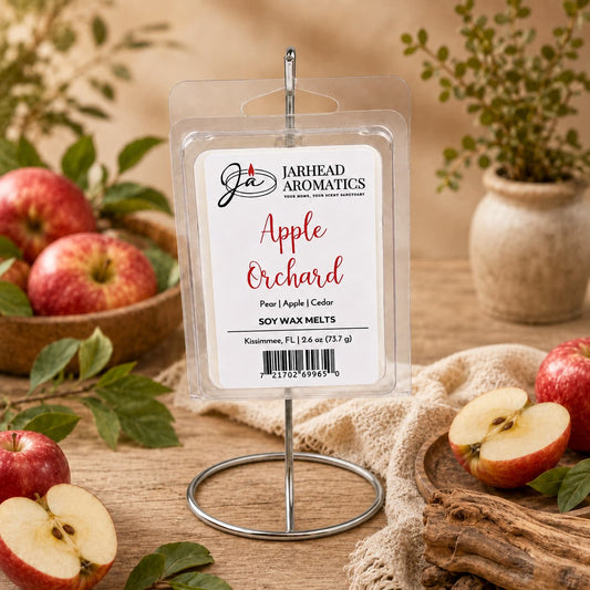 A package of Jarhead Aromatics’ Apple Orchard Soy Wax Melt | Crisp Apple, Juicy Pear & Cedarwood rests on a stand, surrounded by fresh apples, wooden bowls, greenery, and a beige cloth atop a rustic table—capturing the essence of an apple orchard with its long-lasting fragrance.