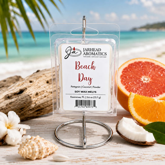 A package of Jarhead Aromatics Beach Day Soy Wax Melt | Citrus, Coconut & Coastal Musk is displayed on a beach surrounded by coconut, orange slices, and seashells, with the ocean and palm trees in the background.