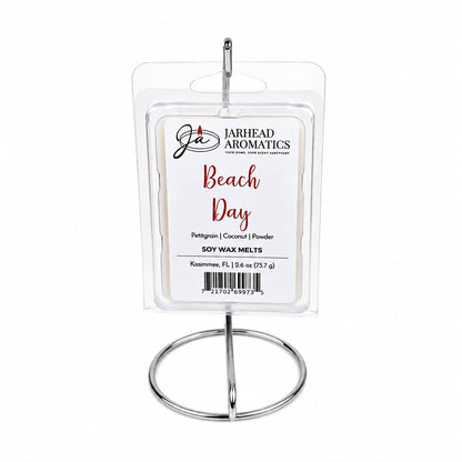 A pack of Jarhead Aromatics Beach Day Soy Wax Melt | Citrus, Coconut & Coastal Musk is displayed upright on a silver metal stand, with the label highlighting its fresh blend of citrus, coconut, and coastal musk scents. This highly fragrant soy wax melt delivers a long-lasting aroma that perfectly captures the essence of a relaxing day at the beach.