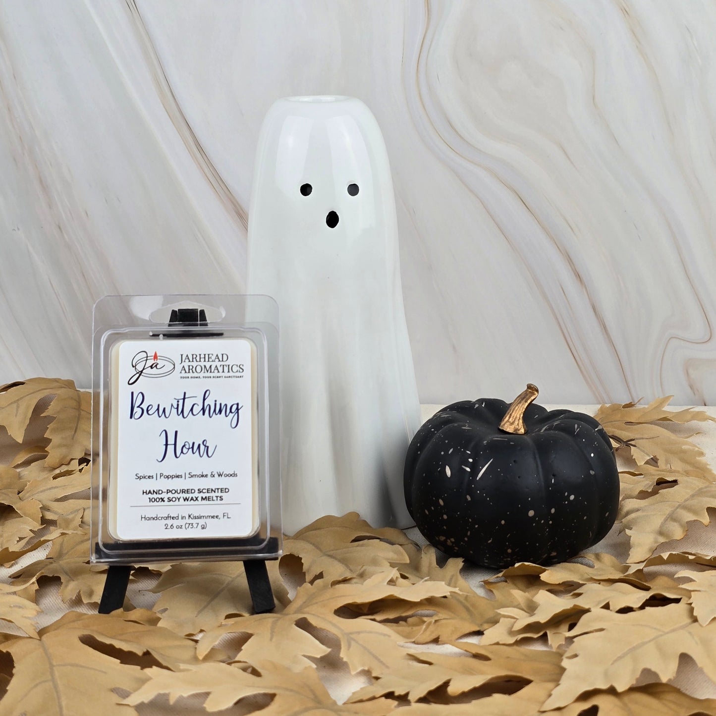 A white ceramic ghost, a black pumpkin, and a Jarhead Aromatics Bewitching Hour Soy Wax Melt from the Halloween Collection with a smoky woods scent sit on tan leaves against a light marble background.