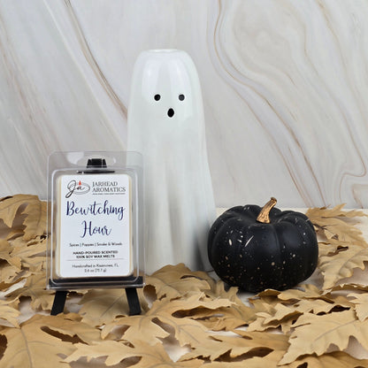 A white ceramic ghost, a black pumpkin, and a Jarhead Aromatics Bewitching Hour Soy Wax Melt from the Halloween Collection with a smoky woods scent sit on tan leaves against a light marble background.