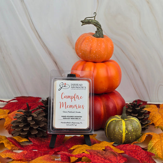 A Jarhead Aromatics Campfire Memories - Soy Wax Melt with a smoky scent is displayed on a small easel, surrounded by pinecones, faux pumpkins, and autumn leaves against a light background.