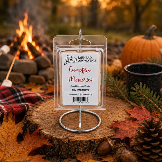 A package of Jarhead Aromatics Campfire Memories Soy Wax Melt | Maple, Clove & Smoky Embers is displayed outdoors on a stand, surrounded by autumn leaves, a pumpkin, a mug, and a campfire in the background. The scene creates a cozy fall atmosphere enhanced by the inviting aroma of maple, clove, and smoky embers—and the long-lasting fragrance of each soy wax melt.