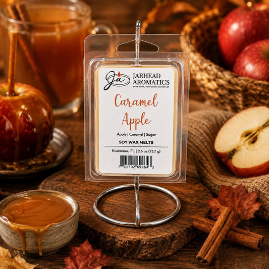A package of Jarhead Aromatics Caramel Apple Soy Wax Melt | Crisp Apple, Rich Caramel & Warm Spice, a bakery-inspired wax melt ideal for fall home fragrance, is displayed on a metal stand, surrounded by apples, caramel, cinnamon sticks, a glass mug, and autumn leaves atop a rustic wooden table.