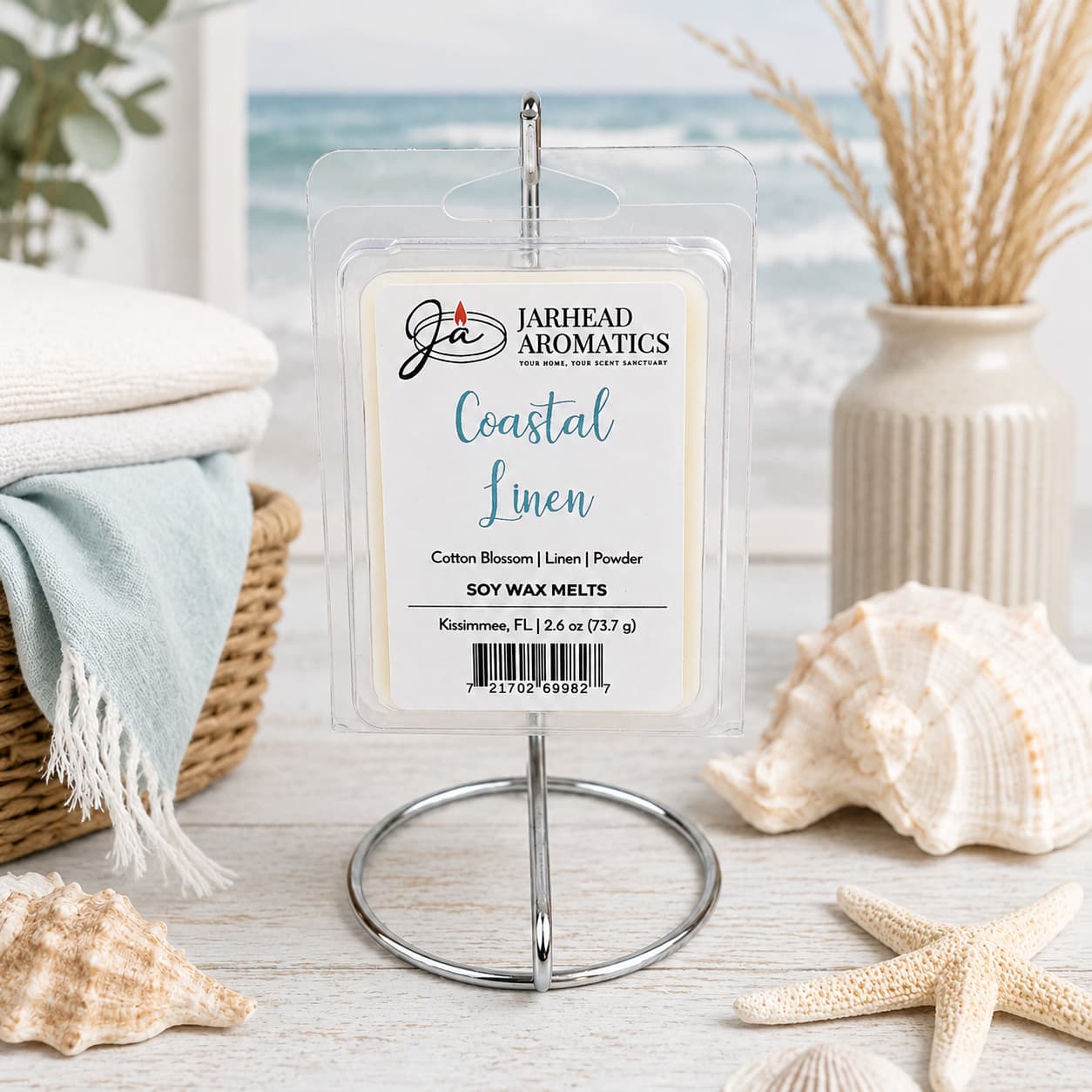 A package of Jarhead Aromatics’ Coastal Linen Soy Wax Melt | Fresh Linen, Ozone & Sandalwood is displayed on a metal stand, surrounded by a seashell, starfish, towels, and beach-themed decor, with a blurred ocean scene in the background. This inviting arrangement perfectly captures the fresh coastal fragrance of this soy wax melt.