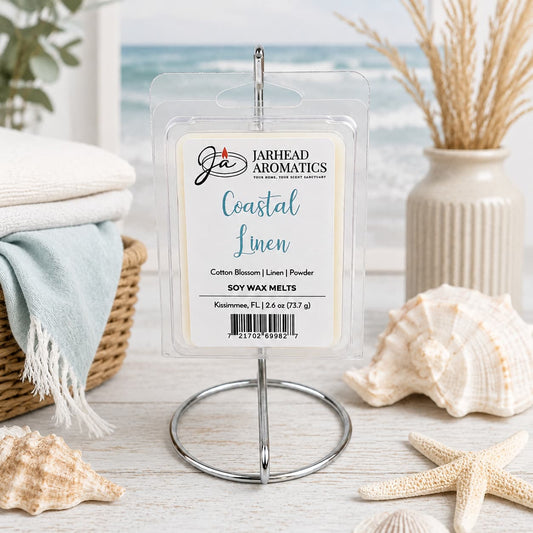 A package of Jarhead Aromatics’ Coastal Linen Soy Wax Melt | Fresh Linen, Ozone & Sandalwood is displayed on a metal stand, surrounded by a seashell, starfish, towels, and beach-themed decor, with a blurred ocean scene in the background. This inviting arrangement perfectly captures the fresh coastal fragrance of this soy wax melt.