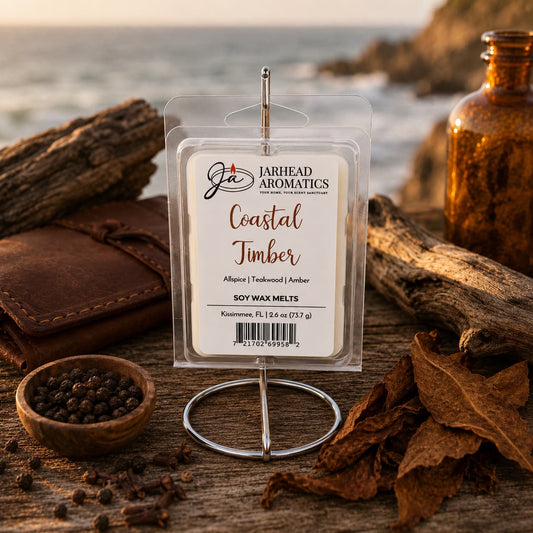 A package of Jarhead Aromatics Coastal Timber Soy Wax Melt | Teakwood, Leather & Warm Amber is displayed on a stand surrounded by spices, wood, and leather, with a blurred ocean and rocky coastline in the background at sunset—a perfect addition to your home fragrance collection.