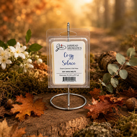 A package of Jarhead Aromatics’ Cozy Solace Soy Wax Melt | Fresh Greens, Jasmine & Oak Moss stands on a metal holder in an autumn forest scene, surrounded by fallen leaves, moss, and white berries. Infused with fresh greens, jasmine, and oak moss, this nature-inspired fragrance offers a long-lasting scent that perfectly captures the tranquility of the season.