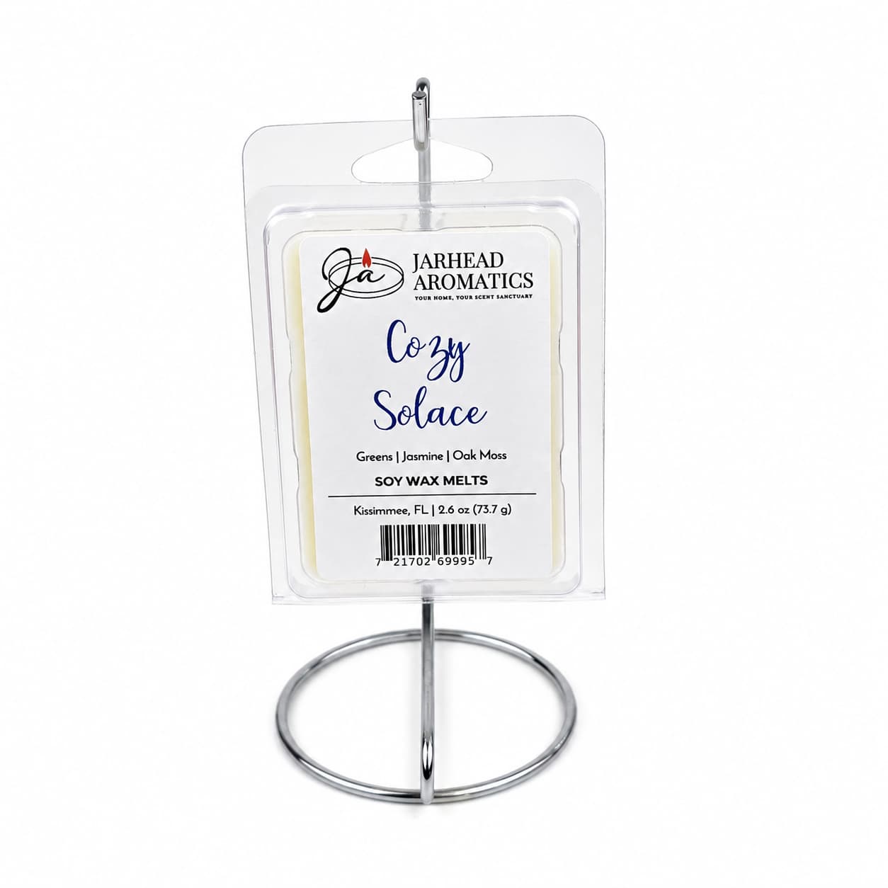 A package of Jarhead Aromatics Cozy Solace Soy Wax Melt | Fresh Greens, Jasmine & Oak Moss is displayed on a silver stand. The label highlights the blend of fresh greens, jasmine, and oak moss by Jarhead Aromatics, offering a nature-inspired fragrance that fills your space with a long-lasting scent.