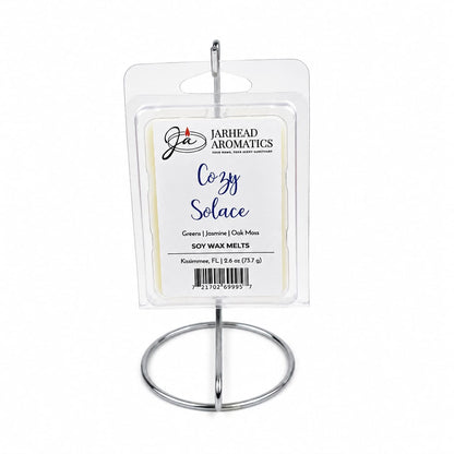 A package of Jarhead Aromatics Cozy Solace Soy Wax Melt | Fresh Greens, Jasmine & Oak Moss is displayed on a silver stand. The label highlights the blend of fresh greens, jasmine, and oak moss by Jarhead Aromatics, offering a nature-inspired fragrance that fills your space with a long-lasting scent.