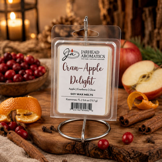 A package of Cran-Apple Delight Soy Wax Melt | Cranberry, Apple & Warm Cinnamon by Jarhead Aromatics is showcased alongside fresh apples, cranberries, cinnamon sticks, and orange peels on a rustic table. The highly fragrant soy wax melt fills the air with its inviting aroma as cozy autumn décor and soft, warm lighting set a festive mood in the background.