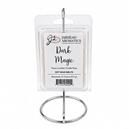 A pack of Jarhead Aromatics Dark Magic Soy Wax Melts, featuring notes of peach, plum, dark florals, and vanilla musk, is elegantly displayed in a clear package on a silver metal stand. The label highlights the enticing fragrance profile of these scented wax melts, with a total weight of 2.6 oz.
