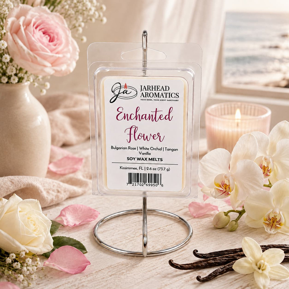 A package of Jarhead Aromatics’ Enchanted Flower Soy Wax Melt, featuring notes of Bulgarian rose, white orchid, and vanilla, is displayed on a stand surrounded by white and pink flowers, a candle, and vanilla beans. A beach scene is visible through the window in the background. The floral fragrance infuses the space with an inviting atmosphere that beautifully complements the serene coastal view.