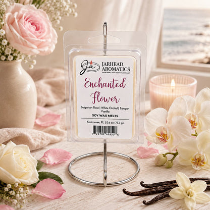 A package of Jarhead Aromatics’ Enchanted Flower Soy Wax Melt, featuring notes of Bulgarian rose, white orchid, and vanilla, is displayed on a stand surrounded by white and pink flowers, a candle, and vanilla beans. A beach scene is visible through the window in the background. The floral fragrance infuses the space with an inviting atmosphere that beautifully complements the serene coastal view.