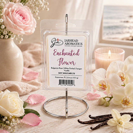 A package of Jarhead Aromatics’ Enchanted Flower Soy Wax Melt, featuring notes of Bulgarian rose, white orchid, and vanilla, is displayed on a stand surrounded by white and pink flowers, a candle, and vanilla beans. A beach scene is visible through the window in the background. The floral fragrance infuses the space with an inviting atmosphere that beautifully complements the serene coastal view.